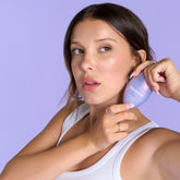 Millie Bobby Brown holding Florence by Mills Glow Forward Glaze Drops near her cheek, posing against a lavender background.