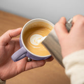 Close-up of hands pouring milk to create latte art in a purple mug.