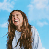 Millie Bobbie Brown laughing joyfully with her eyes closed, set against a bright blue sky with soft clouds.