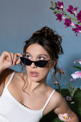 Millie Bobbie Brown wearing black sunglasses and a white textured tank top, glancing to the side with purple flowers in the background.