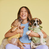 Millie Bobby Brown smiling joyfully while holding two dogs, one in a yellow outfit and the other in green, against a yellow background.